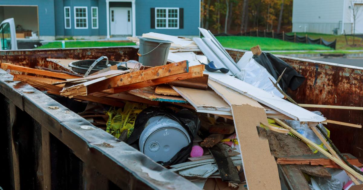 A dumpster filled with wood pieces, plastic buckets, cardboard, and other garbage. Two houses are in the background.