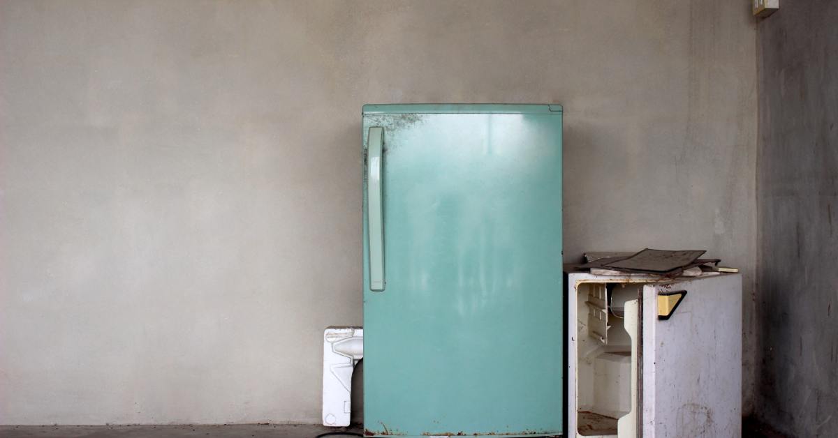 A dark, dingy gray room containing two old refrigerators. One is full-sized, and the other is a mini-fridge with its door ajar.