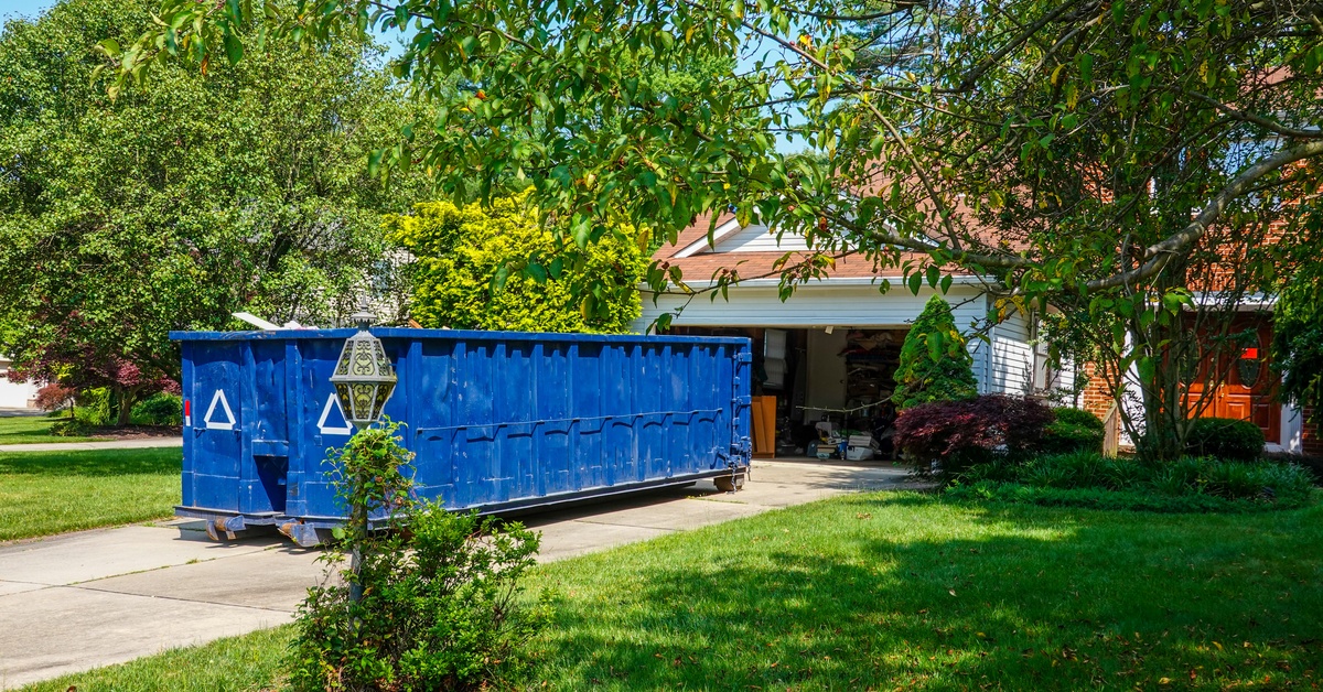 A blue dumpster is parked in the driveway outside the open garage of a home with a lush, green front yard.