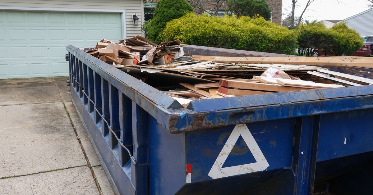 A blue dumpster filled with various construction materials and trash is parked outside a brick home with a garage.