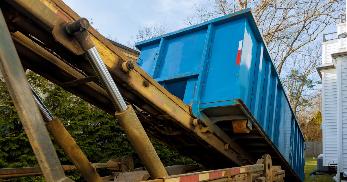 A large, blue roll-off dumpster being loaded off a truck with a hydraulic ramp with a home and yard in the background.