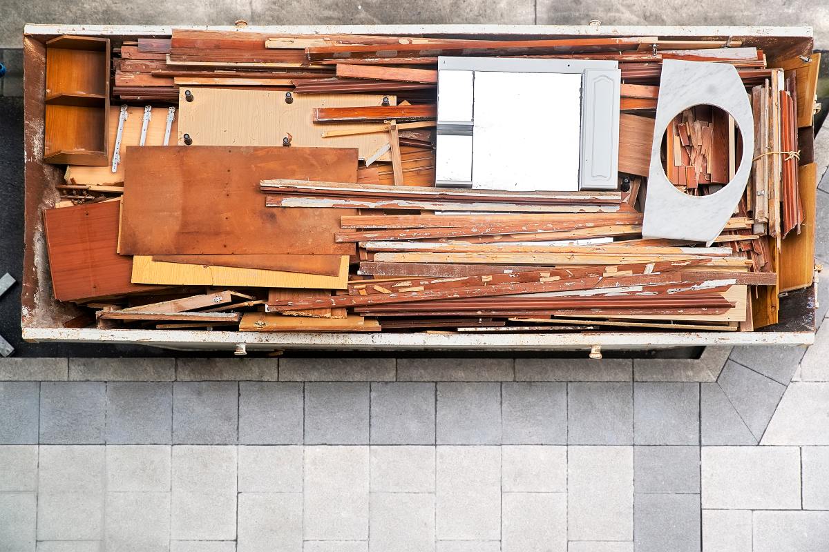 A top-down view of a dumpster at a construction or renovation site, filled with old wood, cabinet, and countertop materials.