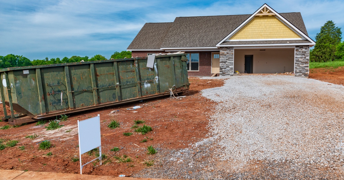 A roll-off dumpster parked in the new yard of a brand-new built home with an open garage and gravel driveway.