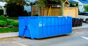 A large, blue roll-off dumpster parked on the curb near a home with a wooden shed in the front yard.