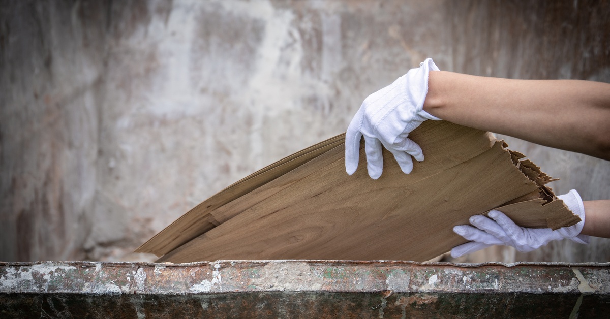 A close-up of a person wearing gloves loading wooden debris into a dumpster with a blurry background.